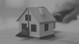 A grayscale image of a small model house, symbolising real estate, home ownership, or architectural design, with a blurred background that suggests a thoughtful or contemplative setting.