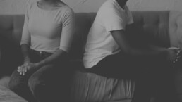 A monochrome image capturing a moment of disconnect, with two individuals seated beside each other on a couch, their body language suggesting emotional distance as they turn slightly away from one another, highlighting a silent story of tension or estrangement.